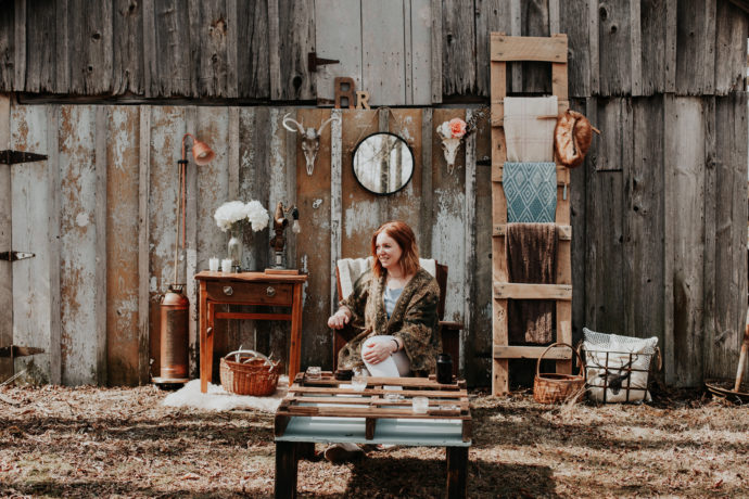 Seated woman in front of a barn door; various home goods, and handmade pieces of furniture are in the background