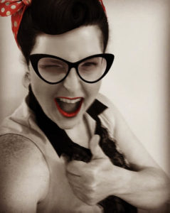 Image description: Theresa, a Deaf Acadian and Indigenous non-binary person, dressed as Adeline, her 1950s style character, wears a bandana on her head, red lipstick, and smiles.