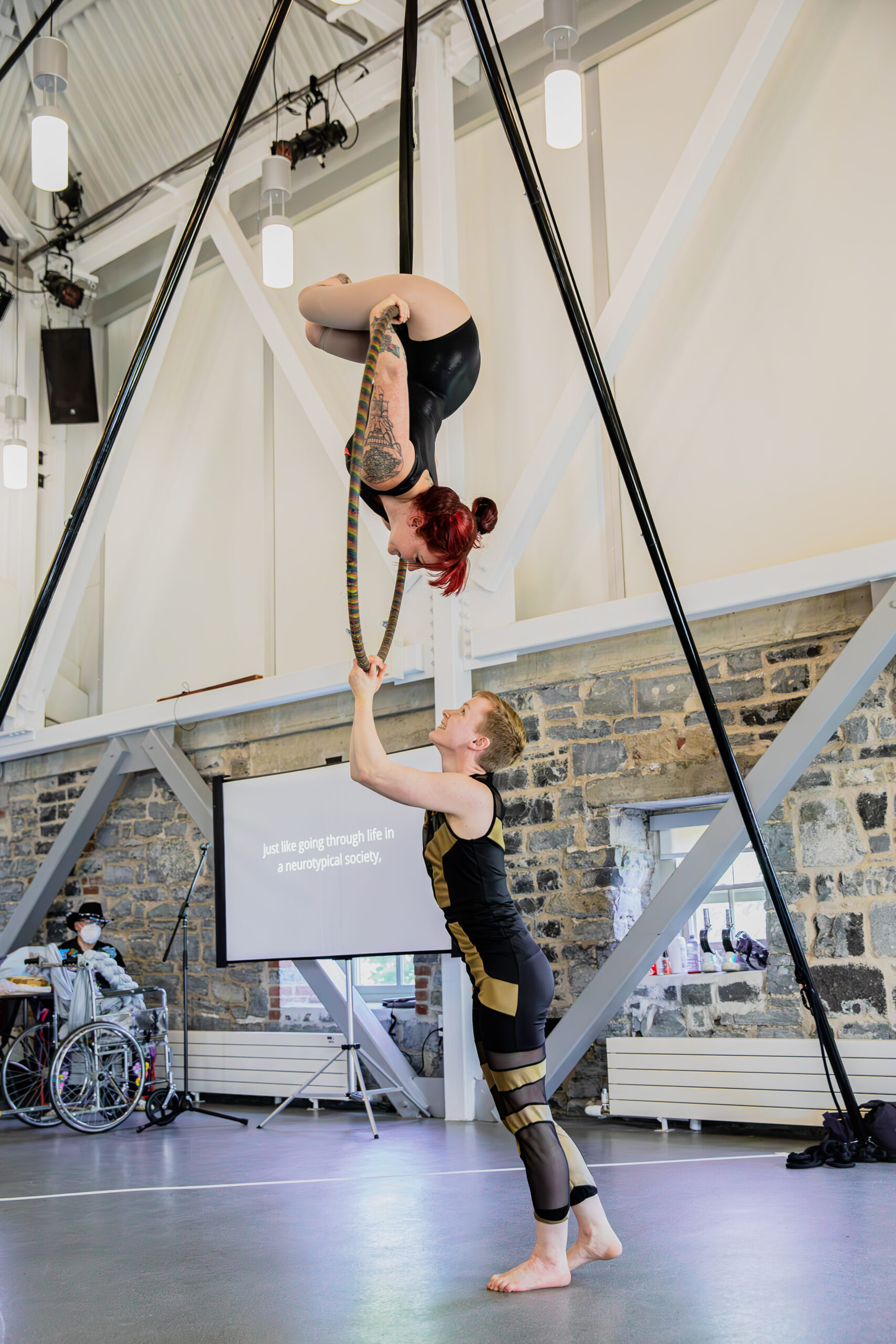A photo of Erin and Maxime during an aerial hoop performance. Erin is a white queer Disabled (double below knee amputee and neuro non-conforming) human with fiery red hair styled with bangs and two buns. Maxime is a white joyful trans and non-binary person with short ginger hair. Erin hangs upside-down from the top of the hoop, with zir body folded over it and both hands holding the hoop. Maxime stands on the ground, with both hands on the bottom of the hoop. Erin and Maxime look at each other joyfully.