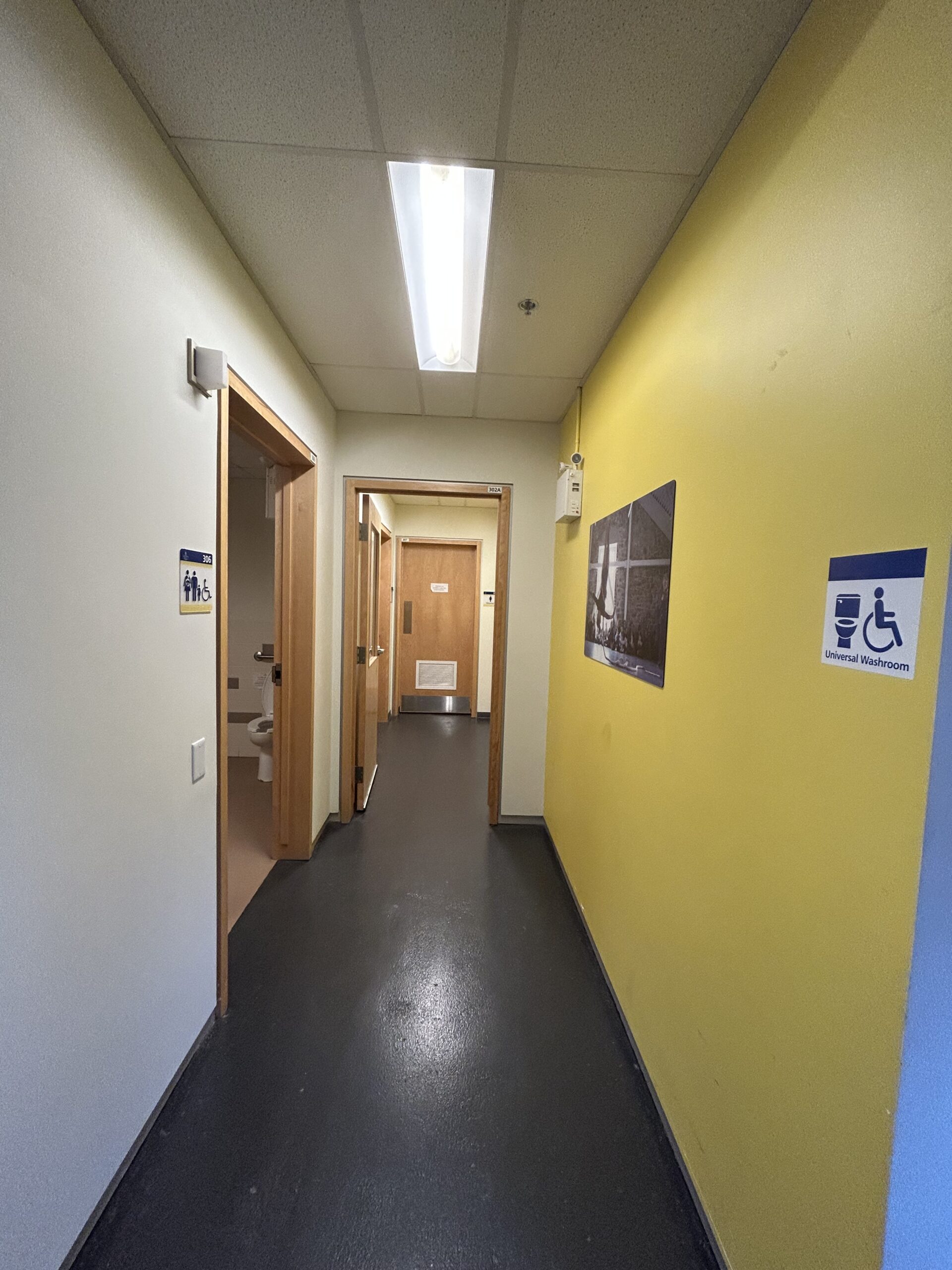 Hallway to washrooms on the third floor [visibility into the universal accessible washroom on the left side of the hallway, propped open door that leads to the entrances for the women's washroom at the end of the hall, the men's washroom is out of the line of sight but is located on the right side]