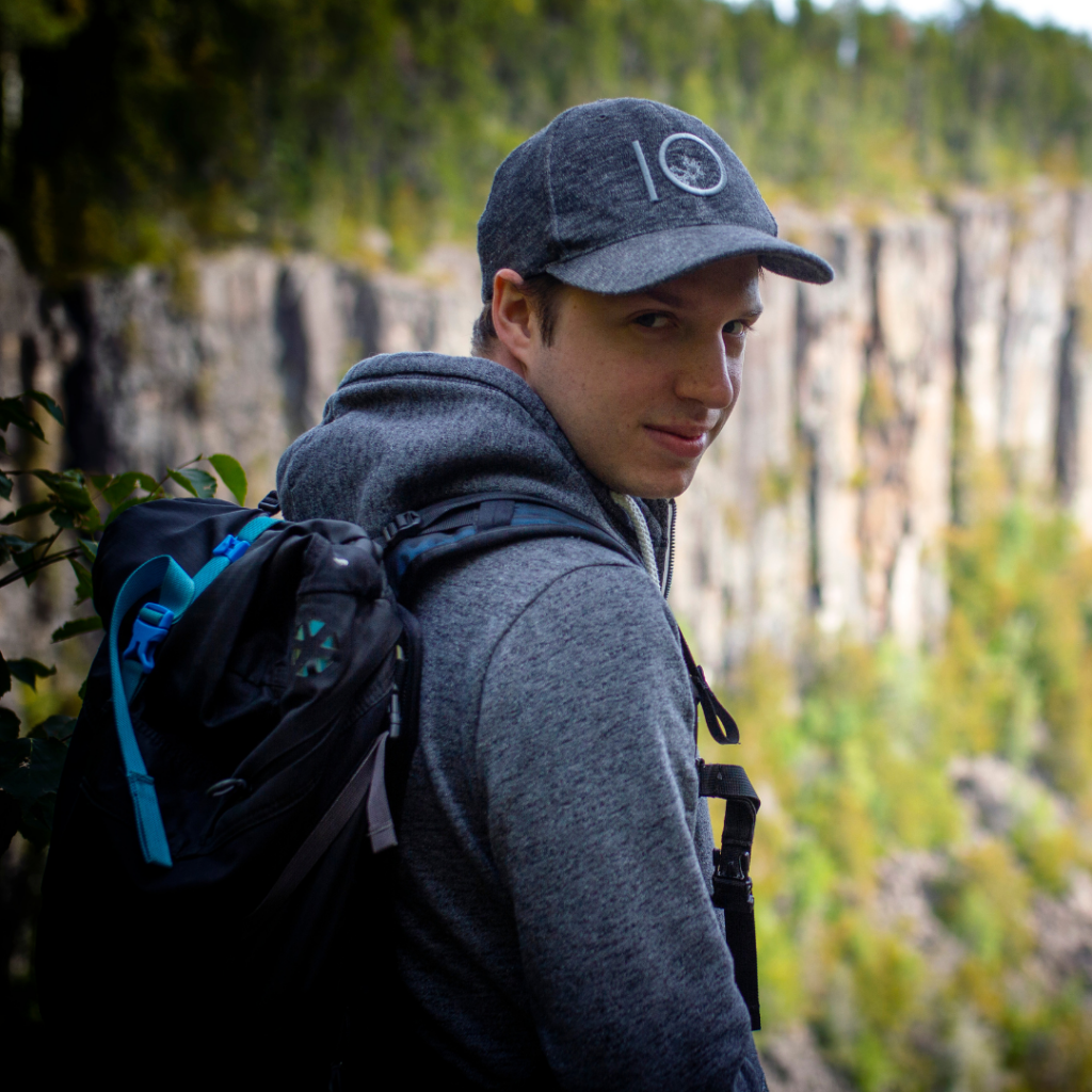 Joe Pagnan stands outside in nature with their back to the camera. They are looking back over their shoulder at the camera. They are wearing a backpack, grey hoodie, and grey 10 tree hat. 